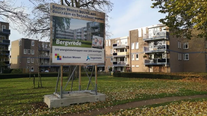 Projectbord op een met herfstbladeren bedekt grasveld bij de Zevenaarse straat Bergvrede met de tekst 'Baston Wonen bouwt aan duurzaam en betaalbaar wonen, Bergvrede. 777 energielabelstappen voor 300 woningen'. Onderaan staan de logo's van Baston Wonen en Hagemans