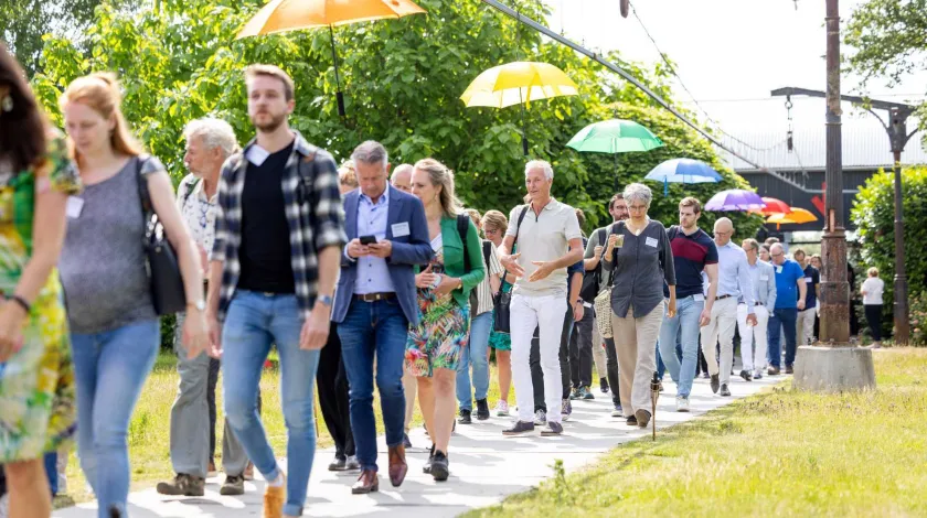 Mensen lopen in een rij over een pad met aan weerszijden gras. Boven hen hangen paraplus in allerlei kleuren.