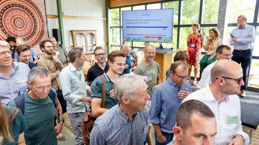 Een groep mensen in een zaal kijken allemaal dezelfde kant op. Op de achtergrond staan drie mensen op een podium.