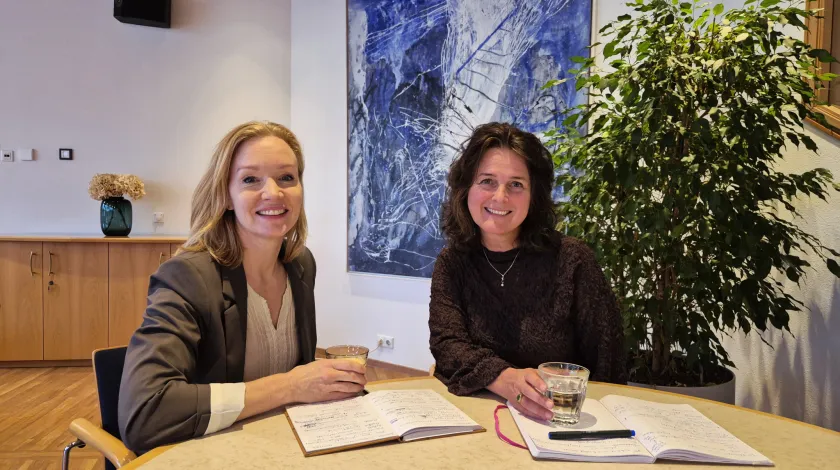 Twee vrouwen aan een tafel met een glaasje water en een schrijfblok voor zich