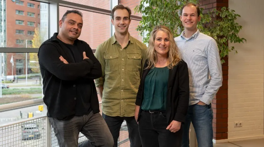 Groepsfoto van 3 mannen en 1 vrouw, die bij een raam staan.