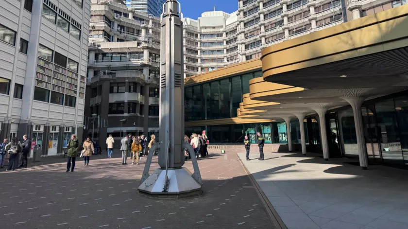 Achterzijde van het gebouw van de Tweede Kamer der Staten Generaal, met een pleintje met een verticaal kunstwerk, voorbijgangers en twee agenten.