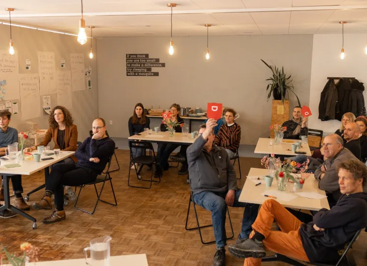 Mensen zitten aan verschillende tafeltjes in een zaal. Twee mannen houden een rood stembordje omhoog.