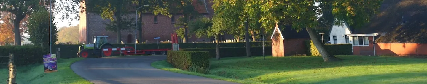 Foto van een weg naar een dorp met rechts een typisch Groningse boerderij, en achter de bomen een kerk