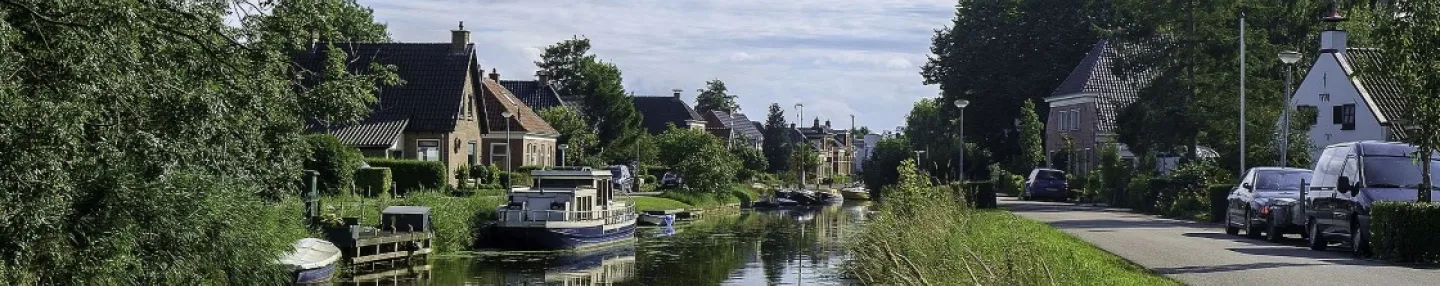 Links een aantal huizen aan een vaart met bootjes aan de steiger, rechts een weg met daarachter een kerktoren