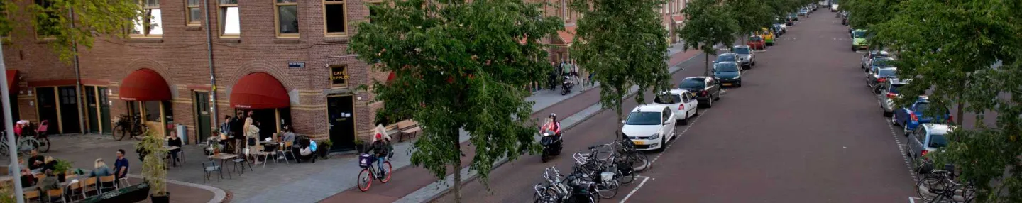 Brede rijbaan met aan weerszijden een rij bomen met daartussen geparkeerde auto's en fietsen. Links Café Keppler met de rode zonneschermen uit en gasten op het terras.