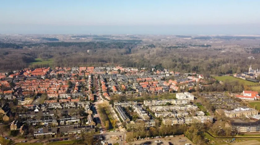 Luchtopname van een woonwijk met oudere en nieuwere laagbouw. Op de achtergrond bossen, duinen en de zee.
