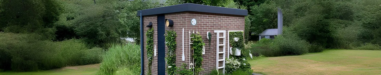Bakstenen transformatorhuisje met begroeiing op een grasveld met op de achtergrond bomen.