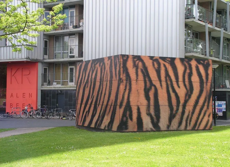 Een trafohuisje in tijgerprint met op de achtergond een galerijflat. Afbeelding genaamd 'Tijger_-_Kralenbeek.JPG' is gemaakt door Richardkw onder licentie van: CC BY-SA 3.0,