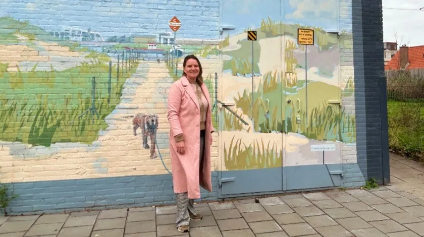 Vrouw in een roze overjas staat voor een transformatorhuisje, dat is beschilderd als een duinlandschap. Aan de rechterkant van het trafohuisje zie je een boom, een elektriciteitsmats en een vak van een villawoning.