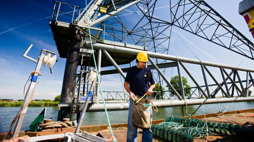 Monteur met gele helm staat op ponton waar een nieuwe elektriciteitsmast op ligt.
