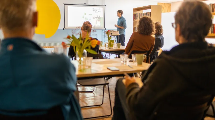 Twee mannen vanaf de rug bekeken luisteren met andere mensen in een lokaal naar een man die een presentatie houdt en daarvoor een projectscherm gebruikt.